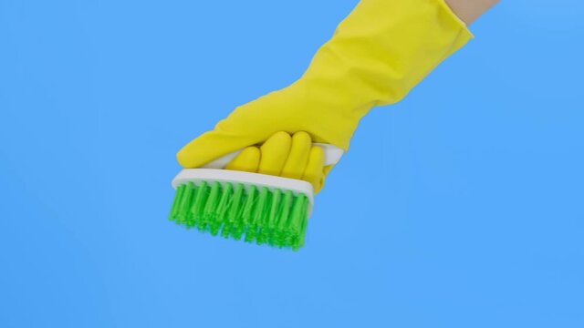 A hand in yellow gloves showing a green brush against blue background, cleaning and brushing carpet, removing stains and wool from it and doing routine homework concept