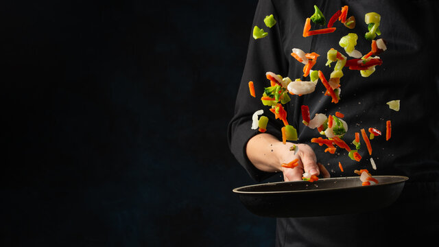 Professional chef in black uniform throws mix of frying vegetables in pan on dark blue background. Frozen motion. Backstage of cooking restaurant dish. Creative preparing meal.