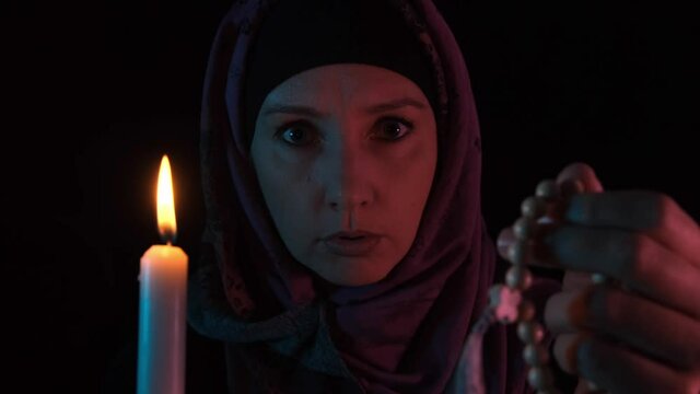A night portrait of a witch casting spell over a candle and rosary, prayer beads and making witchcraft, she is looking at a camera