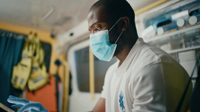 Black African American Paramedic in Face Mask Using Tablet Computer while Riding in an Ambulance Vehicle for an Emergency. Emergency Medical Technicians Outside the Healthcare Hospital.