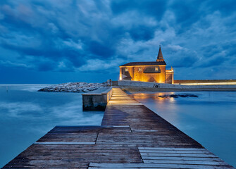 The church of Madonna of the Angel in Caorle, seen from the pier of the east beach at a dark and cloudy sunset at the end of summer (Santuario Madonna dell' Angelo)