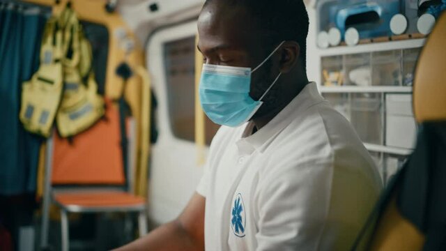 Tired Black African American Paramedic in Face Mask Tries to Rest in an Ambulance Vehicle Going for Emergency. Emergency Medical Technicians are on Their Way to a Call Outside the Healthcare Hospital.