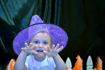 Halloween portrait of little girl wearing purple witch hat scaring the audience against black background. Kid's custom of trick or treat.