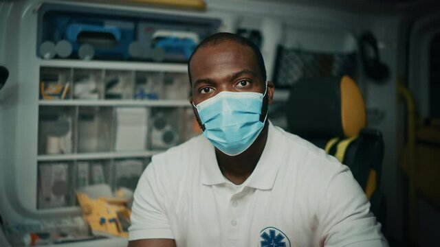 Portrait of a Black African American EMS Paramedic Looks at Camera While Wearing a Safety Face Mask in Ambulance Vehicle. Emergency Medical Technician Outside the Hospital. Covid-19 Concept.