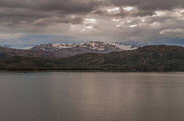 Sarmiento Channel, Chile - December 11, 2008: Amalia Glacier and Fjord. Wide shot of brown cloudscape over snow covered mountain range with belt of green foliage at edge of metalic water.