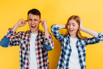 Portrait of young angry people, man and woman covering ears with hands on yellow background