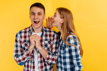 Portrait of young people, man and woman, woman whispering a secret in the ear of a man, gossip on yellow background
