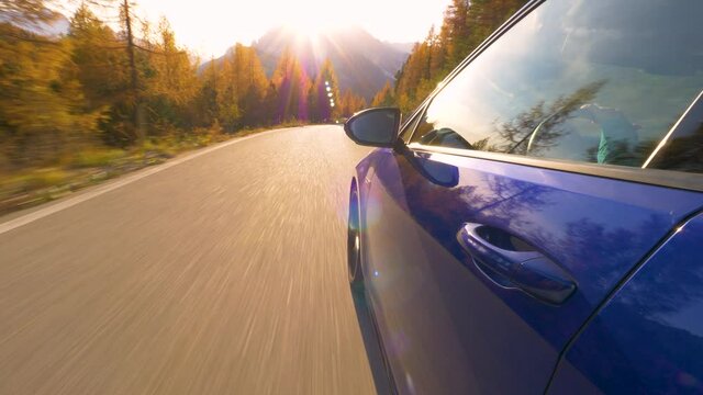 CLOSE UP, LENS FLARE: Golden fall sunbeams shine on a shiny blue car driving along scenic route in the Dolomites. Brand new sportscar speeds down an empty mountain road in the Italian Alps at sunset.