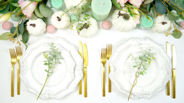 Happy Thanksgiving table setting with white vintage style plates, gold silverware, and centerpiece with greenery, pink, and white pumpkins, on white tablecloth. Top down view overhead styled flat lay.