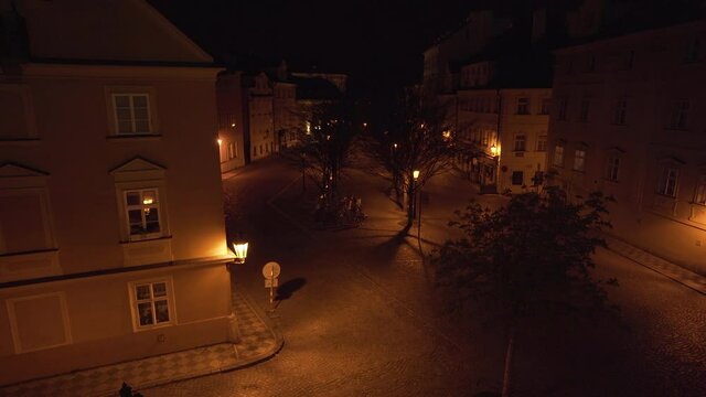 Small empty city square Kampa in Prague, Czechia, with trees and deserted streets during a Covid-19 lockdown at night, houses and pavement lit by golden street lantern light,shot through iron railing.