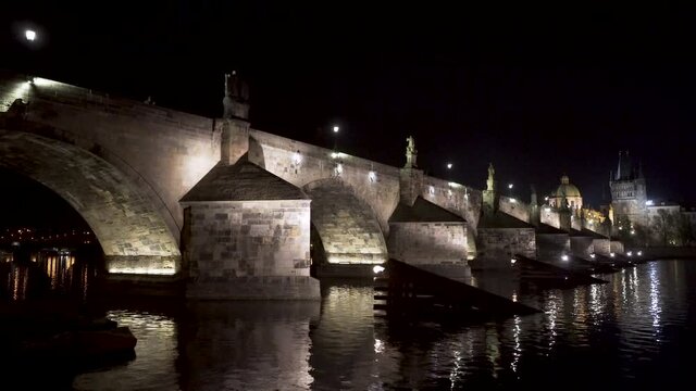 A view of the stone arches of Charles bridge over river Vltava in the historical centre of Prague, Czechia, at night, lit by street lanterns, reflected on the shimmering river surface, pan 4k shot.