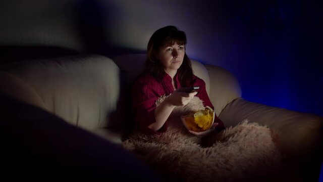 brunette woman sits in sofa with chips in the evening and watches tv with emotion
