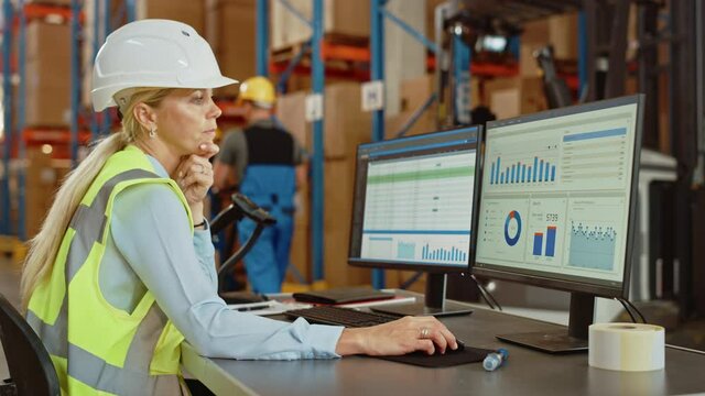 Professional Female Worker Wearing Hard Hat Uses Computer with Inventory Status Checking and Delivery Software in the Retail Warehouse full of Shelves with Goods. Delivery, Distribution Center 