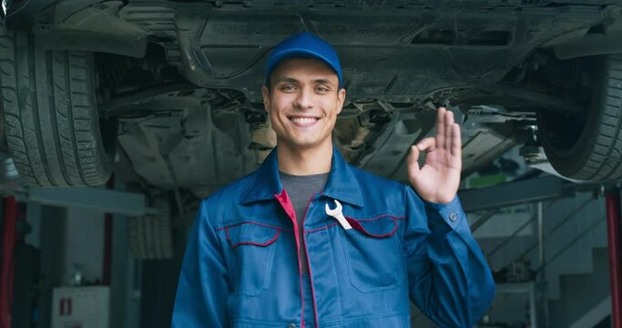Portrait of handsome professional mechanic gesturing OK and smiling to camera, standing at car repair workshop