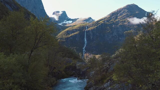 Camera slowly panning through an open forest with a beautiful river and waterfall coming down a mountain in the background. Briksdalen, Norway.