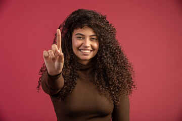 Beauty portrait of a Brazilian woman with afro hairstyle and glamour makeup. Latin woman. Mixed race. Curly hair. Hair style. Red background. Showing One finger.