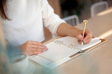 Well groomed woman hand holding gold pen and writing notes with gold pen in notebook beside window.