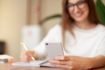 Concentrated young woman writing to do list in notepad and using task management mobile application