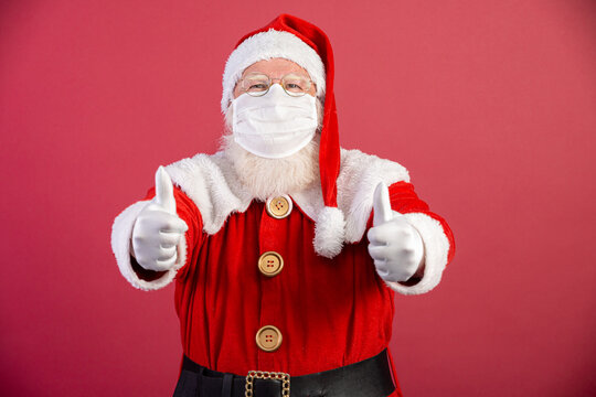 Real Santa Claus with red background, wearing protective mask, glasses and gloves with thumbs up. Christmas with social distance. Covid-19