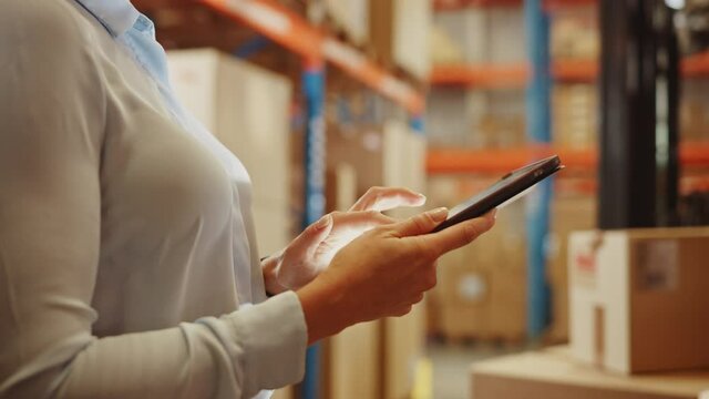 In Warehouse Manager Uses Digital Tablet with App to Check Package Delivery Status with Graphs, Infographics, and Statistics on Screen. Distribution Center with Shelves with Cardboard Boxes 