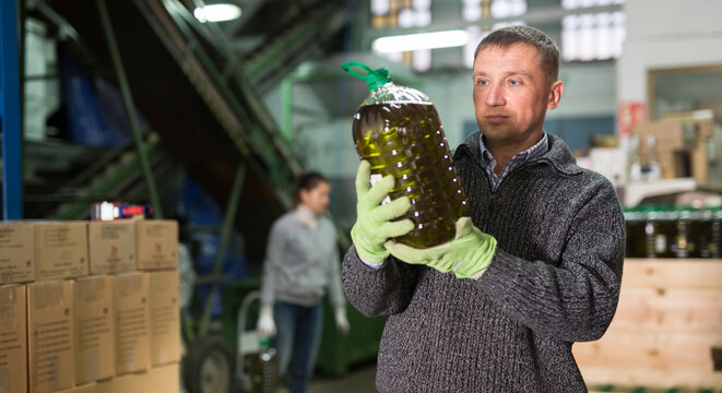 Focused man engaged in family production of olive oil inspecting quality of bottled oil at warehouse - Powered by Adobe