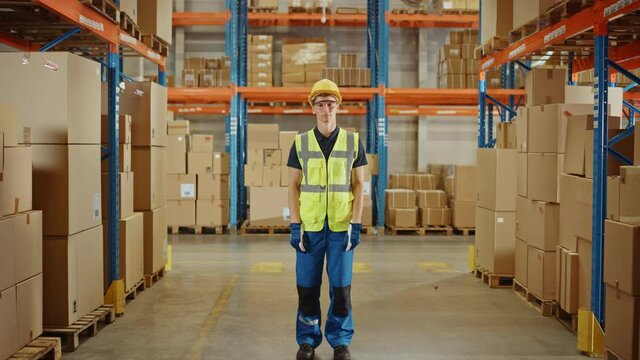 Handsome Smiling Worker Wearing Hard Hat and Safety Goggles Standing in the Retail Warehouse full of Shelves with Cardboard Boxes. Professional Working in Logistics, Delivery, and Distribution Center