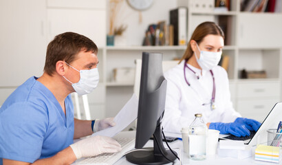 General practitioner wearing medical mask and protective gloves working with case histories on laptop in modern office