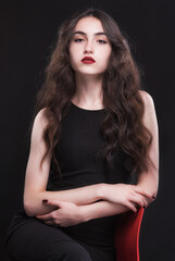Fototapeta premium Beautiful slender young brunette with bright makeup in a black dress on a red chair