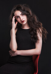 Beautiful slender young brunette with bright makeup in a black dress on a red chair