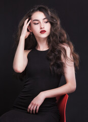 Fototapeta premium Beautiful slender young brunette with bright makeup in a black dress on a red chair