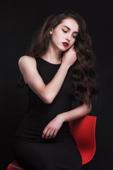 Naklejka premium Beautiful slender young brunette with bright makeup in a black dress on a red chair