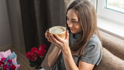 A young beautiful girl is sitting on the bed by the window and enjoying coffee. Concept of a nice holiday morning with a Cup of cappuccino and a good mood