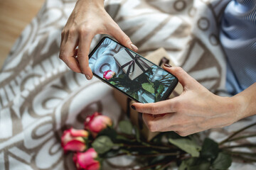 A woman's hands are holding a mobile phone and taking photos of flowers and a gift box on a bedspread. Concept of taking photos on the phone and a birthday or other holiday