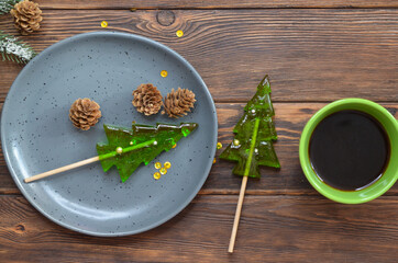 Sweet homemade christmas tree shaped lollipops, children's holiday sweets and dessert, new year's food concept, funny kid's candies, cup of coffee and branch fir or spruce on wooden table