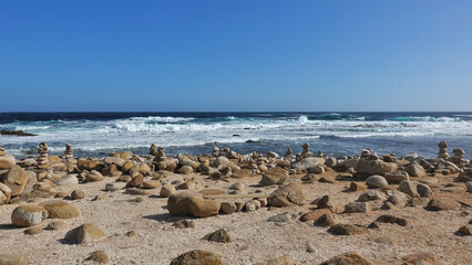 Fototapeta na wymiar The turquoise waves of the Atlantic Ocean roll ashore. White foam. Many rounded stones are stacked in pyramids. On two boulders there is a seating board. Cape of Good Hope. South Africa.