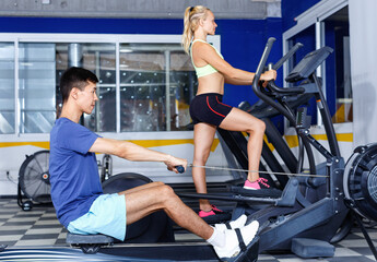 Fototapeta na wymiar Young glad friendly fitness man and woman doing cardio workout on fitness machines at gym
