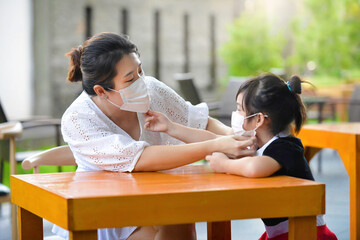 Mother fixing mask on child in outdoor restaurant.Young female and kid with facemask. Social distancing in public.People waiting to be served on the table in bar setting.Pandemic Covid19.