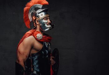 Armoured and serious imperial militray roman posing dressed with steel helmet and red mantle looking away in dark background. - obrazy, fototapety, plakaty