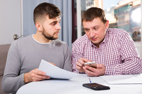 Two serious male friends reading attentively and discussing documents, using phone at home table