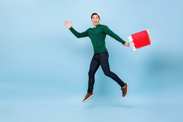 Fototapeta na wymiar Full length portrait of happy young handsome Asian man wearing christmas hat jumping while holding shopping bags isolated on light blue background with copy space