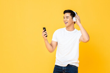 Young handsome Asian man wearing headphones enjoying listening to music from smartphone on yellow...