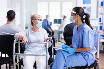 Obraz premium Nurse explaining diagnosis to disabled senior woman with walking frame wearing visor against infection with coronavirus in hospital hallway. Patient and medical staff in waiting area.