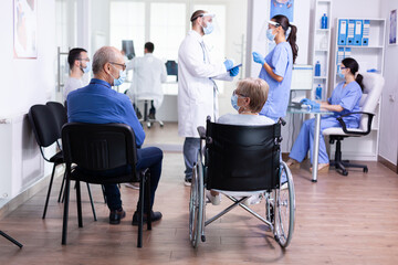 Disabled senior woman wearing face mask against coronavirus in hospital waiting area and having a conversation with old man. Assistant working on reception computer.