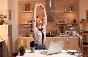 Tired businesswoman streching while working overtime from home kitchen. Employee using modern...