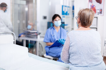 Obraz premium Senior woman wearing face mask against coronavirus outbreak listening nurse during consultation in hospital examination room. Modern private clinic or hospital. Practitioner physician appointment