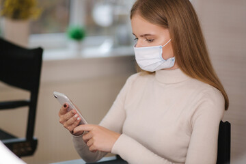 Female customer in beauty salon sitting in medical mask and use phone. Beautiful young blond hair woman wiating for brow master - obrazy, fototapety, plakaty