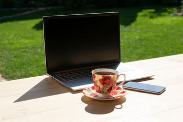 laptop, phone and beautiful porcelain cup of tea stand on light wooden table in green garden. Freelance or distant learning in cozy nice place. Remote work concept.