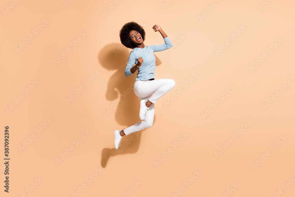 Poster Photo portrait of glad african american woman jumping up holding fists up laughing isolated on pastel beige colored background