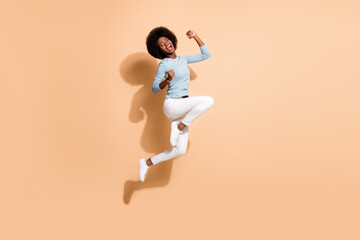 Fototapeta na wymiar Photo portrait of glad african american woman jumping up holding fists up laughing isolated on pastel beige colored background