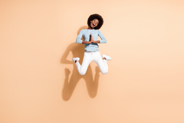 Full length body size photo of curly funny girl with dark skin jumping keeping hands together begging asking isolated on beige color background